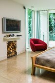 View over bed to red armchair in glazed corner of bedroom, flatscreen TV above wall bracket and stacked firewood in niche