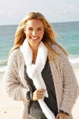 A young blonde woman by the sea wearing a beige knitted cardigan and a scarf