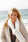 Junge, blonde Frau in beigefarbener Strickjacke und mit Schal am Meer