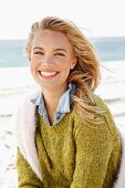 A young blonde woman by the sea wearing a knitted jumper and a denim shirt with another jumper over her shoulders