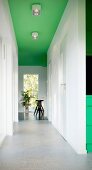 Light-flooded, white corridor with pale grey floor, black bar stool in background and spotlights on green-painted ceiling