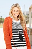 Junge, blonde Frau mit schwarz-weiss gestreiftem Pulli und orangefarbener Strickjacke im Freien