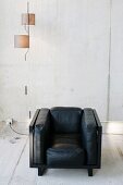 Black, cubic leather armchair and designer pendant lamp with cylindrical lampshades against exposed concrete wall