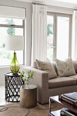 Vase and table lamp on side tables of different heights, ecru sofa and white curtains on terrace doors in background in traditional interior