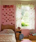 Girl's bedroom in romantic, country-house style with floral bed cover and curtains; wall hanging with heart-shaped pendants on pink wall