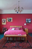 Kronleuchter über Jugendstil Bett und Kleiderbank mit Geflecht; kleine Gemälde auf der himbeerfarben getönten Rückwand und Tagesdecke in Pink