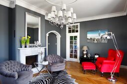 Polstersessel mit grauem Samtbezug vor Kamin und leuchtroter Ohrensessel mit Fussbank in elegantem Wohnraum