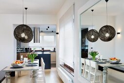 Dining area with wooden table below Moooi pendant lamps with black mesh lampshades in front of open-plan kitchen; large mirror on one wall