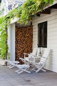 Seating area with white deckchairs and side table below wisteria growing over outside one-storey extension with stacked firewood