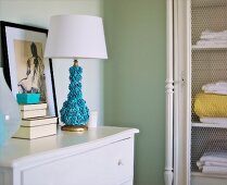 Artistic, turquoise table lamp with floral base on white chest of drawers next to set of boxes and nude drawing in vintage interior