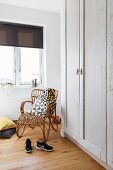 Rattan Sessel im Retro-Stil in Zimmerecke vor Fenster, seitlich Kleiderschrank aus Holz, weiss lasiert
