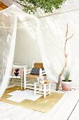 Translucent mosquito net hanging from tree, stool, various chairs and sisal rug on white-painted wooden terrace