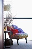 Red cushion on easy chair with zig-zag cover next to vase of twigs on side table and designer lamp