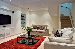 Pale sofas and red rug in front of minimalist wall with fitted cupboards and appliances in living room lit by spotlights