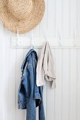 Straw hat and denim jacket hanging from coat pegs on white wood panelling