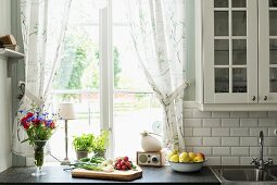 Schneidebrett mit Gemüse, seitlich Blumenstrauss in Vase auf Küchenzeile vor Fenster mit gerafftem Vorhang