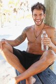 A young tattooed man sitting under a tree wearing shorts and holding a bottle of water