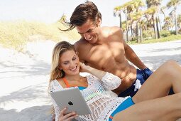 A young couple sitting on a sandy beach with a tablet computer