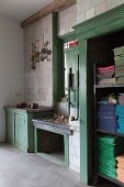 Old, green-painted wooden fittings against tiled wall with decorations hanging from hooks; cloths sorted by colour and stacked in open-fronted cabinet