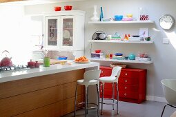 Island counter and white bar stools in open-plan kitchen with colourful crockery on white floating shelves above red chest of drawers