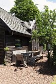 Rustic table, benches and chairs on gravel floor outside wooden house with horizontal exterior cladding