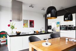 Dining table below black pendant lamps in white, open-plan kitchen with free-standing counter and fitted appliances