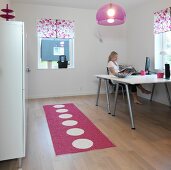 Home-Office mit Doppeltisch und Aktenschrank in schlichtem Weiß, dazu ein gepunkteter Teppichläufer, eine Plexi-Leuchte und Blumenrollos in Pink
