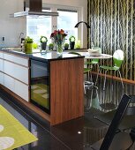 Walnut island counter in front of dining area with green chairs on glossy black-tiled floor and tree-patterned wallpaper