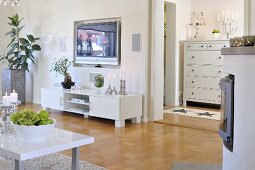 Fruit bowl on coffee table opposite low, white sideboard and flatscreen TV in niche on wall; candlesticks on white chest of drawers in background