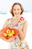 A young woman wearing a summer dress holding a hat filled with fruit and an apple