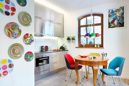 Brightly coloured, retro, upholstered chairs around antique, exotic-wood table, stainless steel kitchen counter and wall plates in open-plan kitchen
