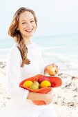 A young woman by the sea wearing a white blouse holding a hat filled with fruit