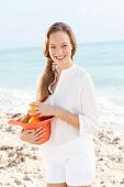 Junge Frau in weisser Bluse und Shorts hält mit Obst gefüllten Hut am Meer