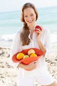 A young woman by the sea wearing a white blouse and shorts holding a hat filled with fruit