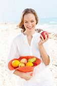 A young woman by the sea wearing a white blouse holding a hat filled with fruit