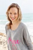 A young blonde woman by the sea wearing a grey jumper with pink writing