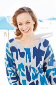 A young blonde woman on the beach wearing a blue patterned jumper