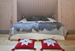 Comfortable bed with grey sheepskin blankets in niche in attic room; red cushions with abstract floral design on wooden floor