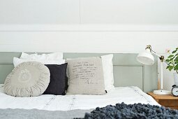 Various scatter cushions in shades of white and grey on bed with grey-painted, wooden headboard