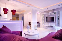 Offener eleganter weißer Wohnbereich mit violetten Farbakzenten, stimmungsvoller indirekter Beleuchtung und Pendelleuchten über Küchentheke