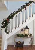 White-painted wooden staircase decorated with fir branches and Christmas decorations above lanterns on console table with curved legs