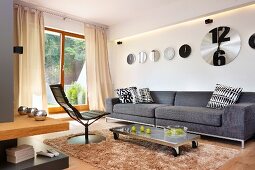 Modern swivel chair and grey couch around coffee table with castors on flokati rug below collection of clocks on wall