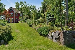 Breiter Rasenweg in sommerlichem Garten, im Hintergrund rotbraun gestrichenes Holzhaus