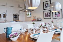 Festively set table below designer pendant lamps with metal lampshades in front of kitchen counter and fitted cabinets