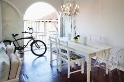 White dining table, refurbished second-hand chairs and Ghost chair below chandelier; bicycle in background in front of open arched terrace doors