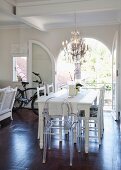 White dining table, refurbished second-hand chairs and Ghost chair below chandelier; bicycle in background in front of open arched terrace doors