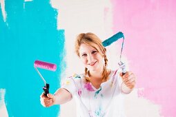 Young woman with hair in plaits holding up two pink and blue paint rollers
