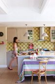 Wooden chair at table with striped tablecloth in front of free-standing kitchen counter in open-plan kitchen; woman in background