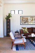 Elegant, curved, 50s armchair on blue and white striped rug and scatter cushion on brown leather sofa in retro interior