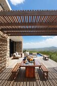 Simple modern outdoor furniture below wooden slatted pergola on spacious terrace adjoining stone house